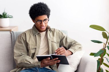 Uzaktan eğitim konsepti. Siyahi üniversite öğrencisi karantina sırasında evden ders çalışıyor. Afrikalı Amerikalı genç sınavı için notlar alıyor, ödev yapıyor ya da online derse hazırlanıyor.