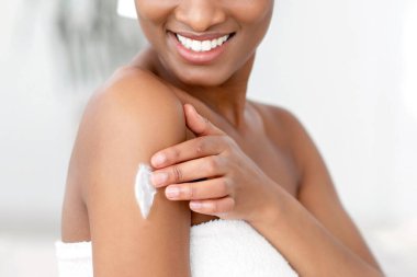 Organik cilt bakımı, rutin ve nemlendirici ürünler. Gülümseyen Afrikalı Amerikalı bayan blogcu beyaz havluyla duş aldıktan sonra elinde krem sürüyor, sabah yatak odasının içinde, boşluğu kopyala