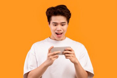 Harika bir uygulama. Portakal rengi arka planda izole edilmiş cep telefonuyla mobil video oyunu oynayan heyecanlı Asyalı gencin portresi, kopyalama alanı. Duygusal adam video izliyor, internette geziniyor.