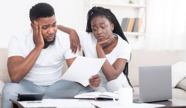 Borç konsepti. Faturaya bakan üzgün afro bir çiftin portresi, büyük faturalara üzülmüş, ev kredisini ödeyemiyor, evdeki kanepede depresif bir şekilde oturuyor.
