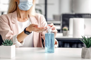 Ofiste güvenlik ve sağlık koruma. Akıllı saatli koruyucu maskeli milenyum kadını iş yerinde antiseptik kullanıyor. El dezenfeksiyonu için dizüstü bilgisayar kullanıyor. Çoklu 19 salgını sırasında.