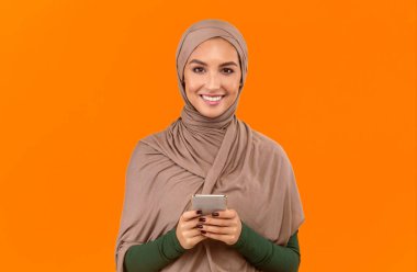 Smartphone kullanan genç müslüman kadın telefondaki yeni mobil uygulamayı onaylıyor. Sarı Stüdyo Arkaplan 'ın başında duruyor, kameraya gülümsüyor. Cep telefonu Reklam Kavramı Uygulaması