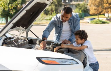Siyah baba ve küçük oğul açık havada araba motorlarını kontrol ediyorlar. Afrikalı Amerikalı baba ve okul çağındaki çocuk araba kaputunun yanında duruyor, bozuk otomobili tamir ediyor, birlikte seyahat ediyorlar.