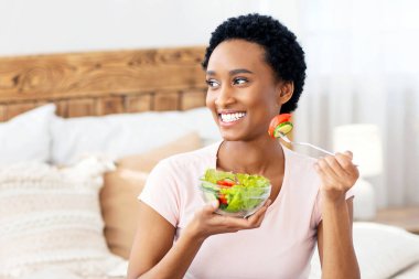 Kilo verme konsepti için sağlıklı diyet. Evdeki yatakta nefis sebze salatası yiyen mutlu siyah kadın, boş alan. Pozitif Afro-Amerikan kadın kahvaltısını dengeliyor.