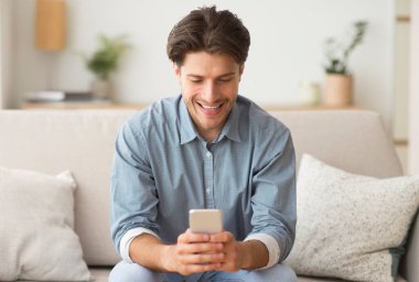 Mobil Uygulamalar. Millennial Guy evdeki koltukta oturan cep telefonuna bakıyor..