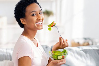 Sağlıklı beslenme konsepti. Güzel siyah kadın evdeki kanepede lezzetli sebze salatası yiyor, panorama. Slim Afrikalı Amerikalı kadın dengeli vejetaryen yemekleriyle kilosunu koruyor.