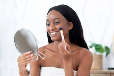 Güzellik hizmetleri için sabah ritüeli. Mutlu Milenyum Çekici Afrikalı Amerikalı kadın duş aldıktan sonra havluya sarılıp yüzüne fırçayla makyaj yapıyor ve yatak odasındaki aynaya bakıyor.