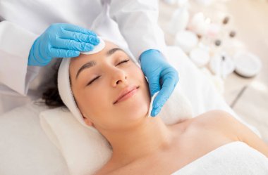 Yüz bakımı. Kozmetik uzmanı pamuk pedleri kullanıyor, salondaki genç Hintli kadının cildini temizliyor, rahat Hindu kadın masaya uzanmış, gözleri kapalı, cilt işleminden keyif alıyor.