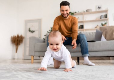 Mutlu genç baba, yeni yürümeye başlayan bebeğiyle oynuyor, babalık izni alıyor, küçük kızıyla evde bağ kuruyor. Çocuk Bakımı, Sevimli Babalık Anları, Ebeveynlik Mutluluğu. Seçici Odaklanma