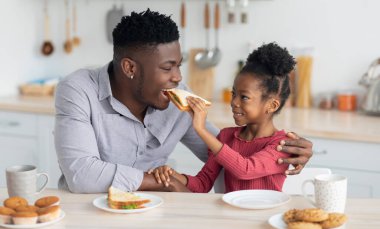 Şirin kıvırcık saçlı, tatlı Afrikalı Amerikalı kız yakışıklı babasını besliyor, ona lezzetli organik sandviç veriyor, siyah baba ve kızı mutfakta birlikte atıştırıyor.