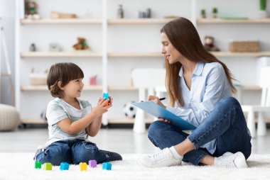 Çocuk psikolojisi sınavı. Renkli plastik bloklarla oynayan sevimli küçük çocuk, genç profesyonel kadın çocuk psikologlar not defterine notlar yazıyorlar, yerde yan yana oturuyorlar.