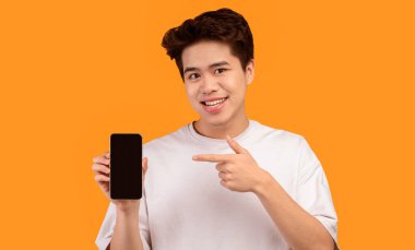 Bu web sitesini tavsiye ediyorum. Elinde siyah boş ekranla akıllı telefonu tutan gülümseyen Asyalı genç bir adam ve cihazı işaret ediyor. Model için boş alanı olan Gadget, turuncu stüdyo arka planında izole edilmiş.
