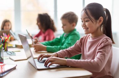 Çevrimiçi Öğrenme. Okuldaki modern sınıflarda dizüstü bilgisayar kullanan çeşitli çocuklar, mutlu Asyalı kız öğrencilerin bilgisayarda internete göz atmasına odaklanıyor. E-Öğrenme, Eğitim ve Teknoloji