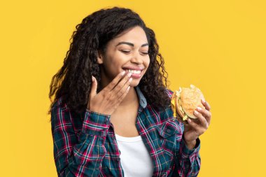 Hile Yemeği. Mutlu Afro-Amerikalı Kadın Lezzetli Burger yiyor, gülüyor ve dudaklarını parmaklarıyla siliyor, boşluğu kopyalıyor. Fast food 'u elinde tutan tatmin olmuş kadın, turuncu arka planda izole edilmiş, afiş.