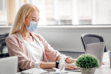 Yeni normal, modern ofis virüsten ve müdürlük işinden korunuyor. Koruyucu maskeli sarışın bir iş kadını. Bilgisayarda antiseptik şişesiyle yazı yazıyor. Karantinadan sonra çalışıyor.