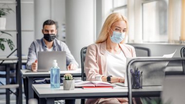 Yeni çalışma koşulları ve karantinadan sonra sosyal mesafeler. Koruyucu maskeli sarışın kadın iş yerinde evrak ve dizüstü bilgisayarla çalışıyor. İç tarafında da büyük bir şişe antiseptik var.