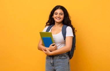 Eğitim Konsepti. Pozitif gülümseyen Hintli kız öğrenci elinde defterlerle kameraya bakıyor, reklam için fotokopi çekiyor. Sırt çantalı kadın, sarı stüdyo geçmişi var.