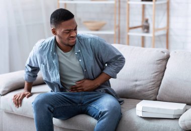 Afro-amerikalı adam pizza yedikten sonra karnına dokunduktan sonra evde koltukta oturmaktan acı çekiyor. Gıda zehirlenmesi, mide iltihabı, karın iltihabı ve apandisit..