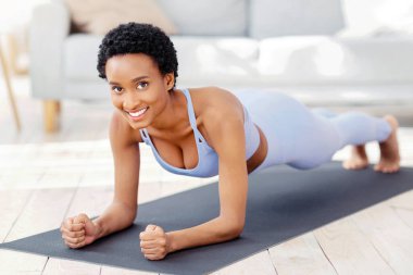 Spor kıyafetli gülümseyen zenci bir kadının uzun bir portresi. Evdeki spor minderinde dirsek tahtası yapıyor. Formda bir Afrikalı Amerikalı kadın ev içi eğitim alıyor, yoga yapıyor ya da evde pilates yapıyor.