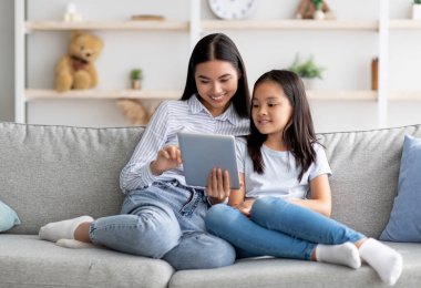Mutlu Asyalı anne ve kız dijital tablet kullanıyor, video izliyor ya da internette sörf yapıyor, evdeki kanepede oturuyor, kopya çekiyor. Genç Koreli aile birlikte ped kullanıyor.