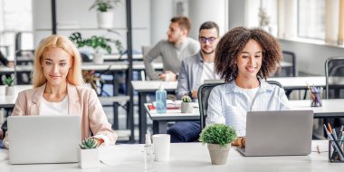 Modern ofiste çalışmak, dijital teknoloji ve karantinadan sonra sosyal mesafeler. Avrupalı ve Afro-Amerikalı gülümseyen kadınlar dizüstü bilgisayarlarda çalışıyor, içleri antiseptiklerle dolu, boş bir masada oturuyor.