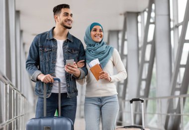 Havalimanında bavul, pasaport ve akıllı telefonla dikilen neşeli İslami eşlerin portresi, mutlu Müslüman çift, tesettürde birlikte seyahat eden Arap erkek ve kadın, seyahate hazır, kopyalama alanı