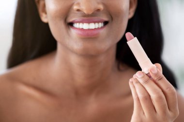 Dudaklarla ilgilenmek, deriyi nemlendirmek ve kozmetik ürünler. Gülümseyen bin yıllık Afro-Amerikan kadın güzellik blogcusu ruj tutuyor ve aynaya bakıyor, blog için video çekiyor, kırpılmış, panorama