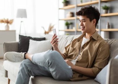 Havalı Gadget ve Uygulama. Cep telefonu tutan, mesaj yazan, oturma odasındaki kanepede oturan Asyalı genç adamın portresi. İnternette gezinen adam, internette sörf yapan, uygulamayı kullanan, boş alan