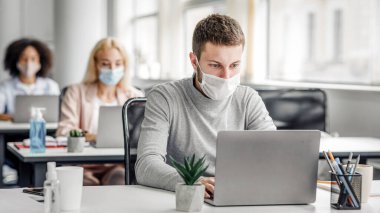 Coronavirus salgını sırasında birlikte çalışma ve güvenli çalışma. Yüzü maskeli genç adam tecritten sonra işe geri döndü ve laptopa baktı, çok ırklı iş arkadaşları gibi, serbest alan gibi.