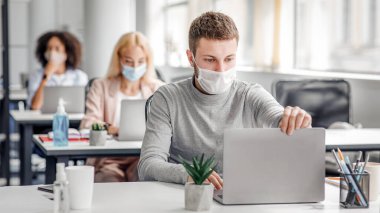 Coronavirus karantina ve ofis işleri sosyal mesafeyi koruyan iş arkadaşlarıyla. Koruyucu maskeli genç adamın portresi masadaki antiseptik laptoplu müşteriyle video görüşmesi yapıyor.