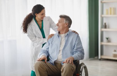 Tekerlekli sandalyedeki mutlu yaşlı adam ve huzurevindeki kadın hemşiresinin portresi. Özürlü, olgun bir erkek hasta. Bakıcısı poz veriyor ve gülümsüyor. Kapalı alanlarda birbirlerine bakıyor.