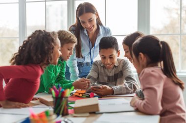 Multiethnic School Kids And Teacher Using Smartphone And Educational Application Learning Sitting At Desk In Modern Classroom At School. Education And Gadgets Concept. Selective Focus