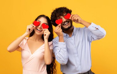 Lovers blinded by their big love. Young indian couple in love holding red heart-shaped cards over eyes and smiling, standing over yellow studio background. St. Valentines Day celebration concept