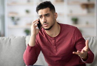 Üzgün Arap adamın kanepede oturup cep telefonuyla konuştuğu yakın plan. Sinirli ortadoğulu genç adam akıllı telefondan konuşuyor, kız arkadaşı ya da müşteri hizmetleriyle kavga ediyor.