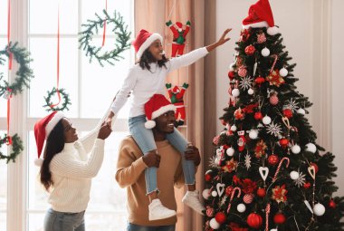 Mutlu aile dekorasyonları Noel ağacı. Kızını omuzlarında tutan genç siyah adam Noel Baba şapkasını üzerine asan Noel Baba ağacını süslemeye yardım ediyor, insanlar oturma odasında birlikte duruyorlar.