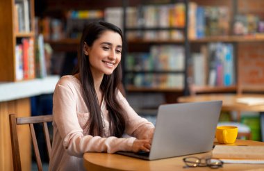 Uzaktan öğrenme ve online çalışma konsepti. Mutlu Arap kadın rahat şehir kafesinde dizüstü bilgisayar kullanıyor, boş yer var. Milenyum Latin bayanı serbest çalışıyor, web konferansına katılıyor.