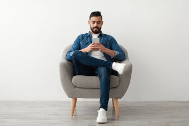 Yakışıklı genç bir Arap cep telefonu kullanıyor, internette sohbet ediyor, koltukta oturuyor, sosyal medyada beyaz stüdyo duvarına karşı sörf yapıyor. Uzaklık iletişimi, günlük tutma kavramı