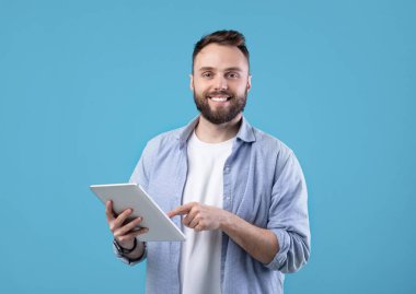 Modern teknolojiler ve iletişim. Mavi stüdyo geçmişi üzerinde tablet bilgisayar kullanan mutlu sakallı adamın portresi. Dokunmatik bloknotlarda çalışan ya da okuyan genç adam.