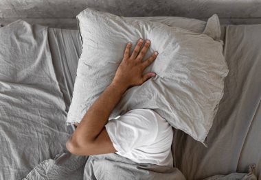 Closeup of irritated young black man lying in bed and covering head and ears with pillow, hearing and suffering from too loud sound. Angry millennial guy cant sleep, tired of noisy neighbors