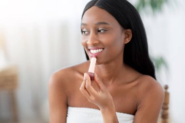 Kozmetik, güzel dudaklar ve kış bakımı. Milenyum Afro-Amerikan gülümseyen kadın kameraya bakıyor, matte ruj tutuyor, ev içinde rutin makyaj yapıyor, yakın çekim yapıyor.