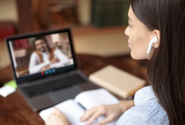 Çevrimiçi eğitim, e-öğrenme kavramı. Kulaklık takan genç bir kadın, erkek öğretmenle video konferansı yapıyor, masada oturuyor ve dizüstü bilgisayar kullanıyor, notlar alıyor, öğrenim materyallerini tartışıyor, ev içini.