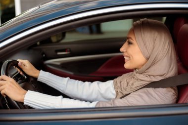 Test sürüşü konsepti. Yeni lüks araba kullanan tesettürlü müslüman kadının yan görünüş portresi. Türbanlı neşeli Arap kadın sürücü koltuğunda oturuyor, direksiyon başında.