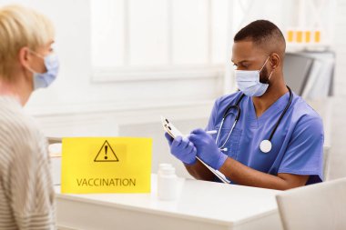 Covid-19 Aşılama. Siyah doktor Coronavirus için hasta aşısı yaptırıyor, hastanede oturup notlar alıyor. Corona Virüs Aşısı, Kapsamlı Önleme. Seçici Odaklanma