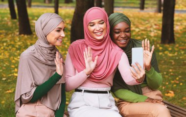 Şehir parkındaki bankta akıllı telefona el sallayan üç Müslüman kadın videosu. İslami Kadın Arkadaşları Video Görüşmesi Yapıyor. E-Öğrenme, Modern Uzaktan İletişim Konsepti
