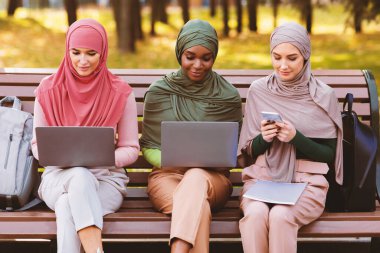 Modern Arap Öğrenci Kadınlar Akıllı Telefon ve Laptop Bilgisayar Kullanıyor Çevrimiçi Bilgisayar Öğreniyor Açık Park 'ta yan yana oturuyorlar. E-Öğrenme, Uzaktan Eğitim Konsepti