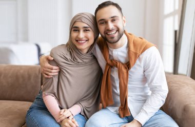 Kameraya gülümseyen mutlu Müslüman çift evdeki koltukta birlikte oturuyor. Hijab 'da Güzel Eşe Sarılan Arap Kocayı Sevmek. Aile, Aşk ve İlişki Konsepti