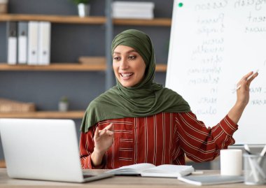 Online Eğitim Konsepti. Beyaz tahtaya işaret eden tesettürlü gülümseyen Arapça öğretmeni portresi öğrencilere İngilizce dil bilgisi kurallarını açıklıyor. Heyecanlı kadın dizüstü bilgisayara bakıyor, konuşuyor.