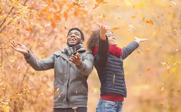 Afro-Amerikalı çift sonbahar parkında yapraklarla oynayarak eğleniyor.