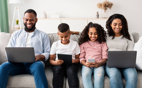 Family Gadgets. Portrait of happy African American parents and their little children holding and using different electronic devices while sitting on the couch in living room at home