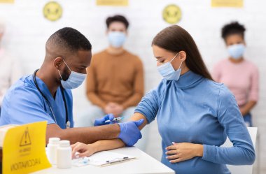 Covid-19 Aşılama. Hastane odasında oturan hamile kadına Coronavirus aşısı yapan siyah bir doktor. Covid 19 Nüfus Bağışıklık Kampanyası, Corona Virüs İlaç ve Tedavi